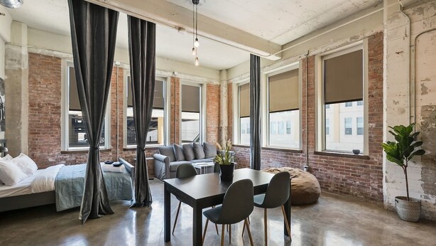 A modern, industrial-style room with exposed brick walls, large windows, a sofa, a dining table with chairs, and a bed. - Beautiful 3bd Unit in Perfect Downtown Location (Dallas)