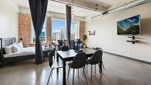 A modern hotel room with a bed, a dining table with chairs, a TV mounted on the wall, and a large window with curtains. - Downtown Dallas Gorgeous 3 BD, Pool & Gym (Dallas)