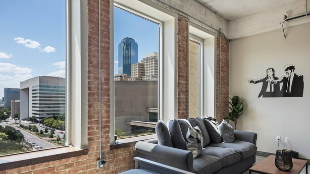 A modern living room with a large window, a grey sofa, and a city view. - Downtown Dallas Gorgeous 3 BD, Pool & Gym (Dallas)