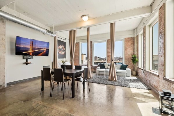 A spacious living area with a dining table, chairs, and a sofa. There is a large window with curtains, a TV mounted on the wall, and a plant on the floor. - 3 Bedroom Unit in Downtown Dallas with Pool & Gym (Dallas)