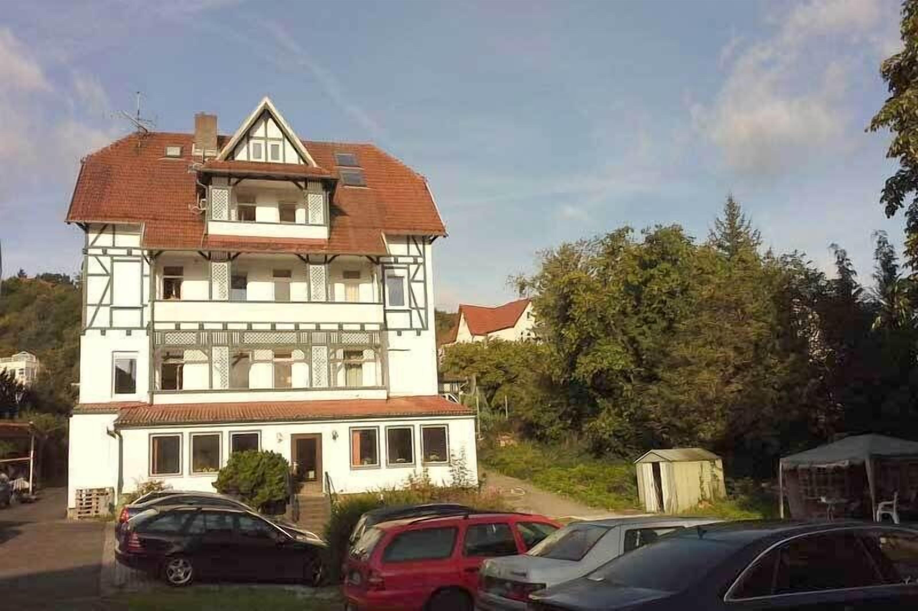 Un edificio de dos pisos con techo a dos aguas y un balcón en el nivel superior. Se ven varios coches estacionados al frente, y una pequeña caseta es visible a la derecha.