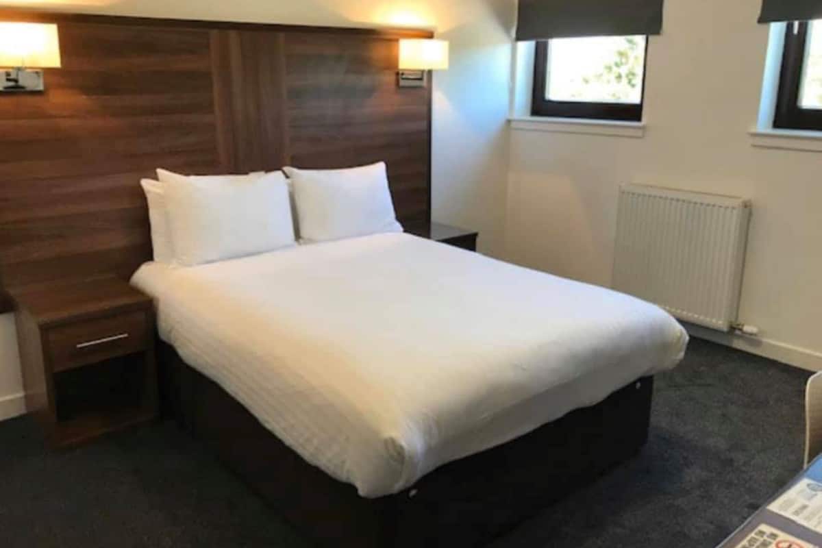 A hotel room with a wooden headboard, two bedside lamps, a white bedspread, a nightstand, a window with a view, and a radiator.