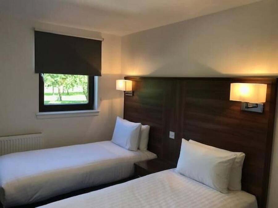 Habitación de hotel con dos camas individuales, ventana con vista a zonas verdes y lámparas fijadas a la pared.