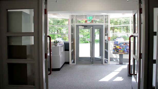 Un área de recepción con puerta de vidrio, mostrador de recepción y un tablón de anuncios con diversos avisos.