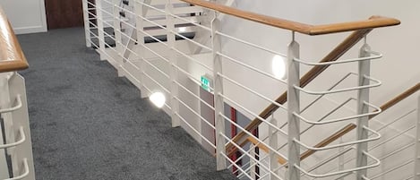 A modern staircase with a wooden handrail and metal railings, carpeted floor, and a door labeled 'Rooms 127-132'.