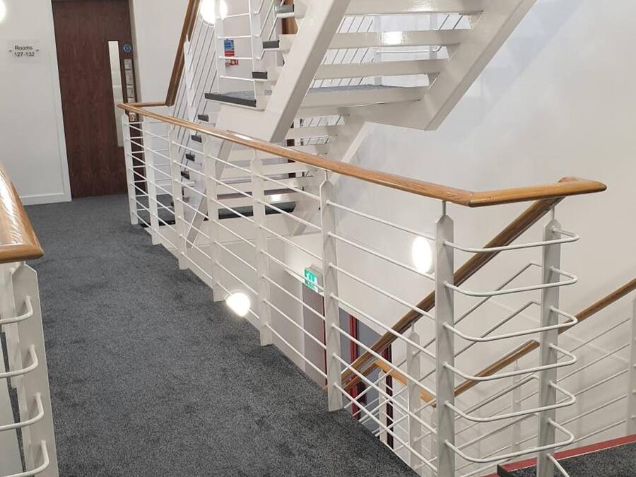 A modern staircase with a wooden handrail and metal railings, carpeted floor, and a door labeled 'Rooms 127-132'.