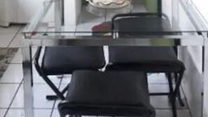 Uma cozinha compacta com uma mesa de vidro e duas cadeiras pretas, uma geladeira branca e uma porta verde que dá para outro quarto.
