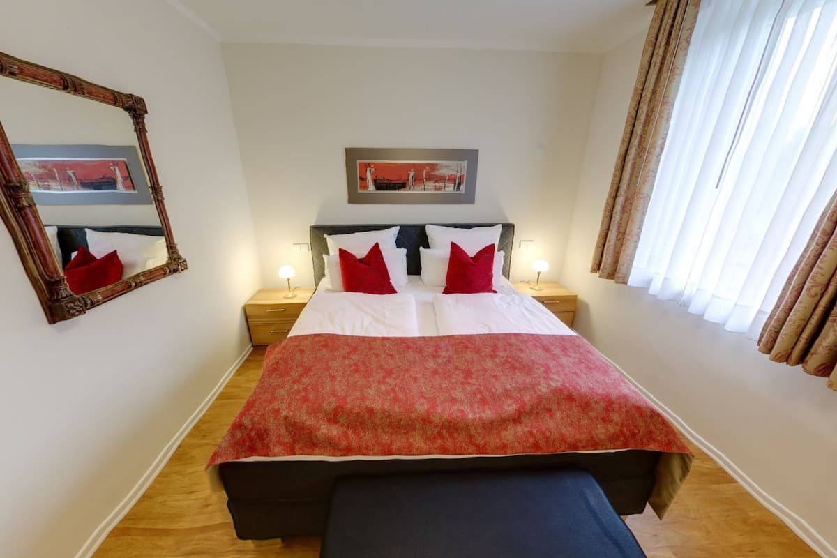 A neatly made bed with a red and white bedspread, two bedside lamps, a large mirror, and a window with curtains.