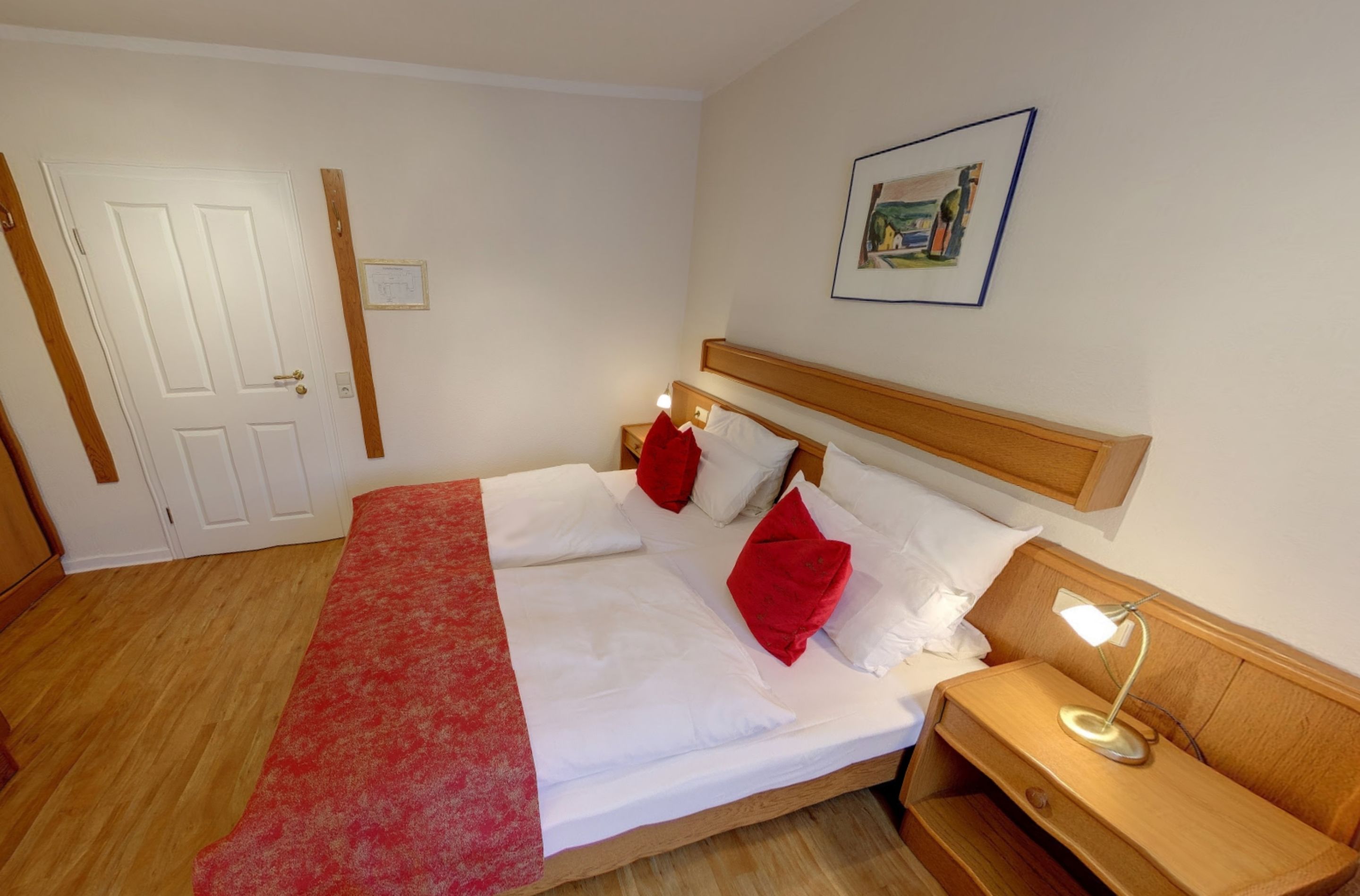 Una cama bien hecha con colcha roja y blanca, cabecera de madera y mesita de noche con lámpara.