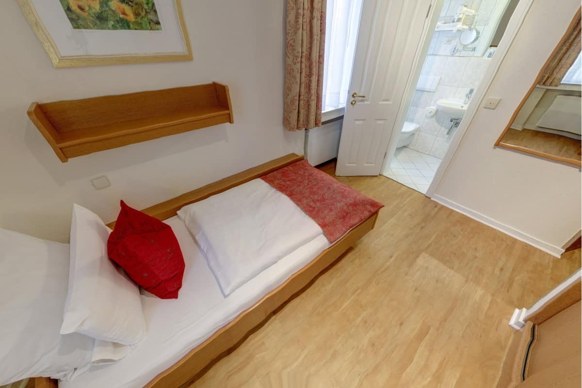 A small, neatly made bed with white and red bedding, a wooden headboard, and a small shelf above it.