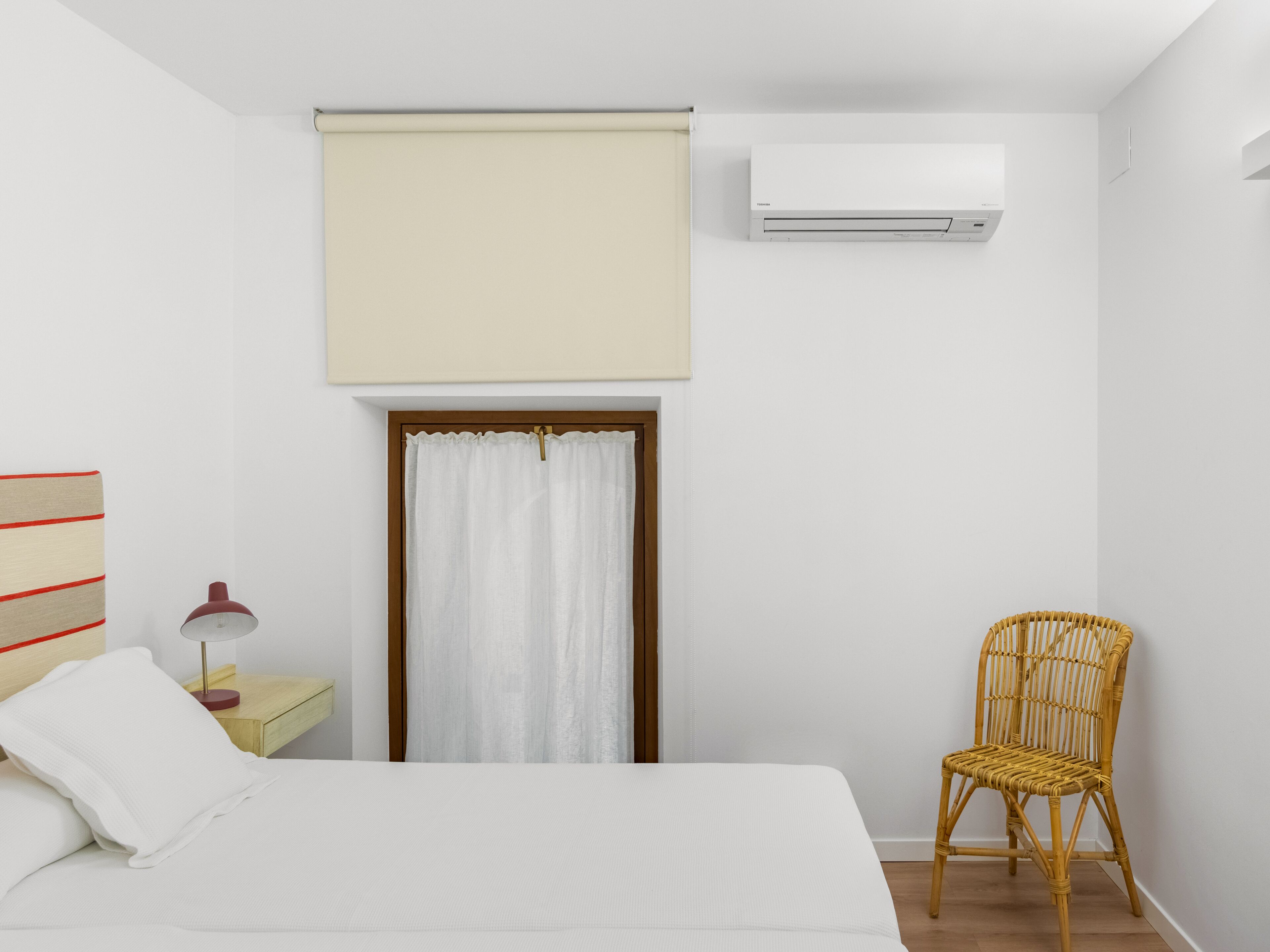 Een slaapkamer met een bed, een stoel, een nachtkastje, een airconditioner en een raam met gordijn.