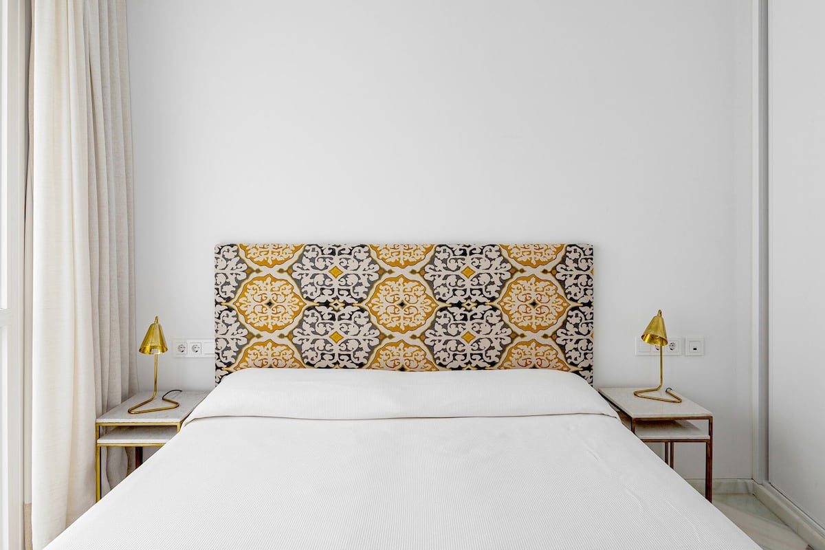 A bedroom with a patterned headboard, a bed with a white cover, two bedside tables with lamps, and a window with curtains.