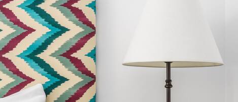 Ein Bett mit weißer Bettwäsche, ein farbenfroher Kopfteil mit Zickzackmuster, ein Nachttisch mit einer Lampe und ein Paar Hausschuhe.