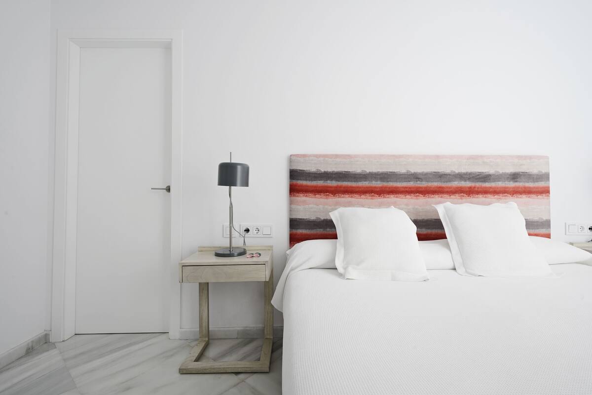 A hotel room with a bed, a bedside table with a lamp, and a wall with a striped artwork.