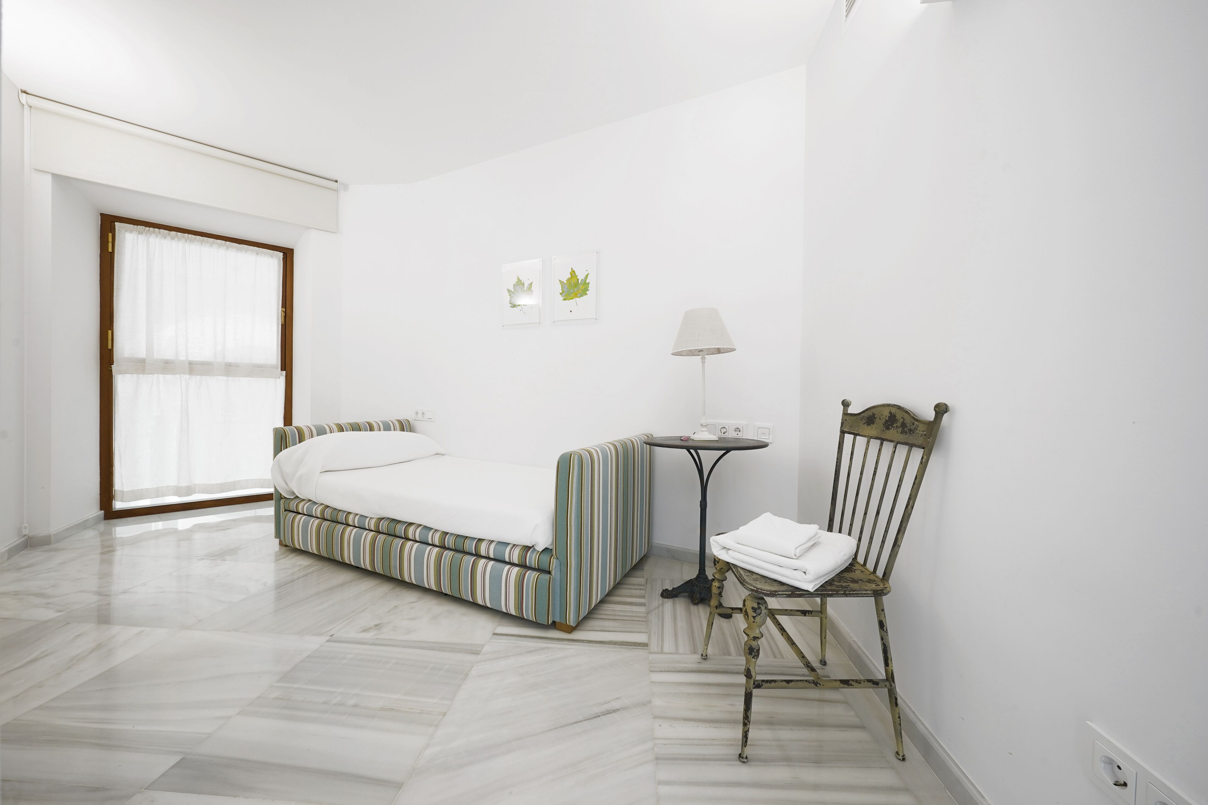 Un dormitorio con una cama, una silla, una mesita y un ventanal.