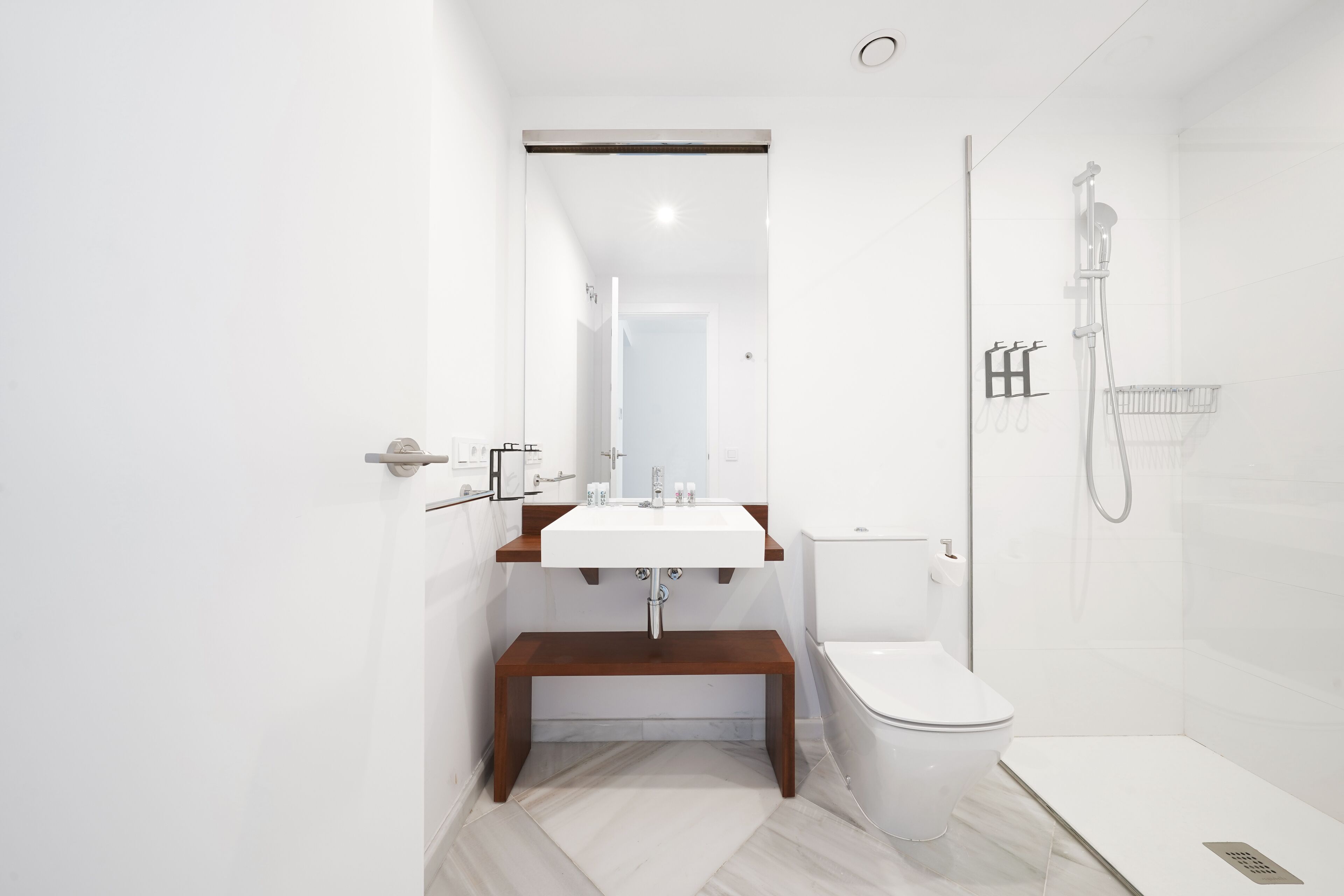 Un baño moderno con lavamanos blanco, banco de madera, inodoro y zona de ducha con cerramiento de vidrio.
