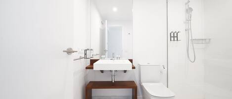 Un baño moderno con lavamanos blanco, banco de madera, inodoro y zona de ducha con cerramiento de vidrio.