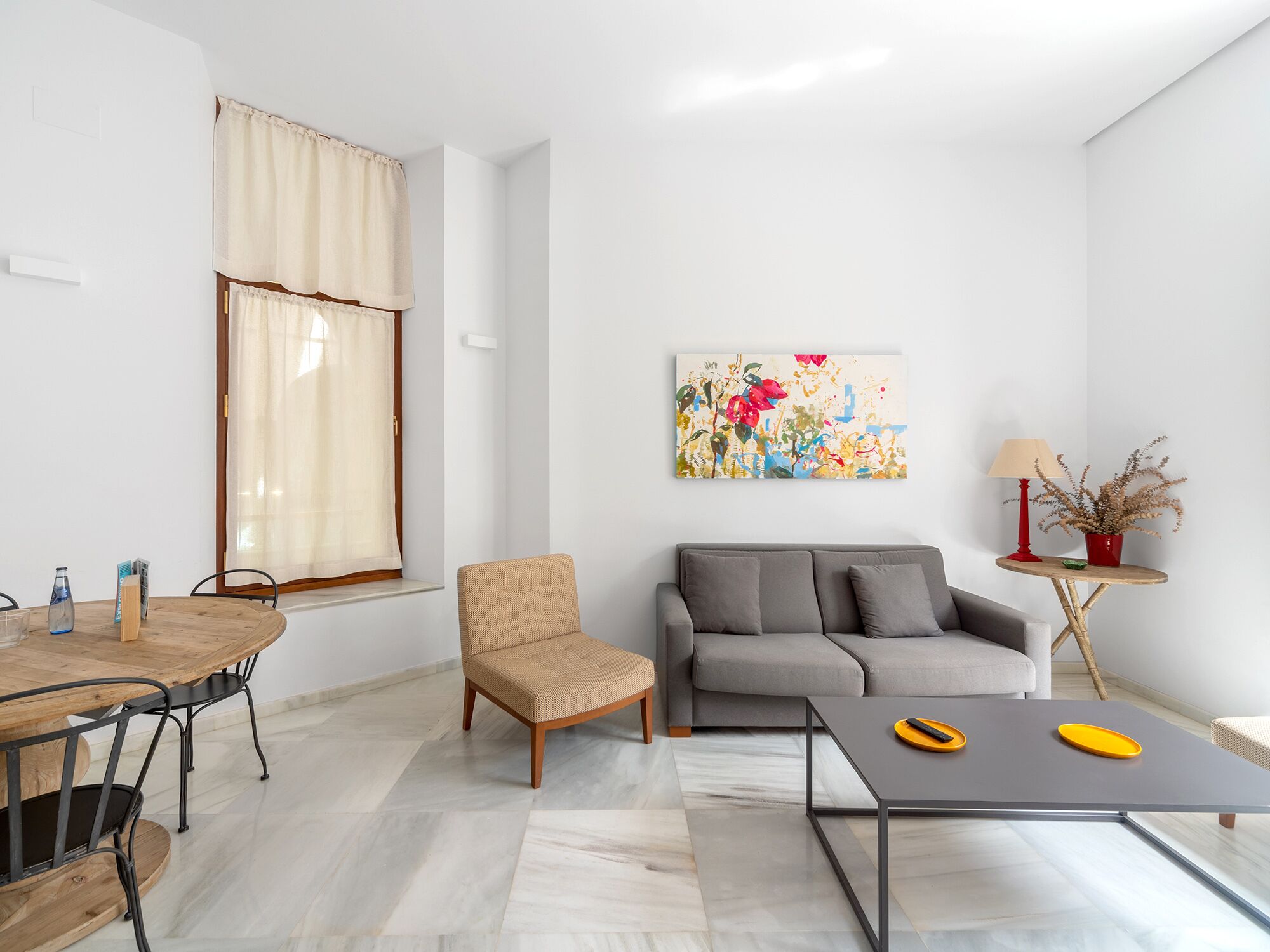 A modern living room with a sofa, a coffee table, a dining table with chairs, a window with curtains, a painting on the wall, and a small side table with a lamp and a plant.