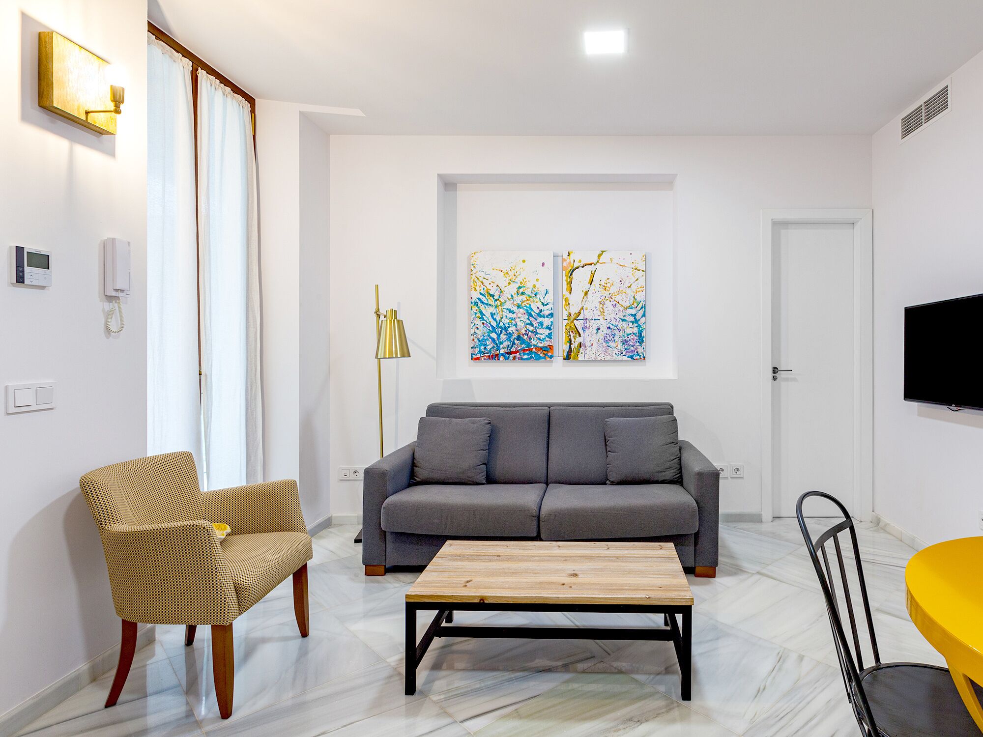Ein modernes Wohnzimmer mit einem grauen Sofa, einem Holztisch, einem Korbstuhl und einem gelben Sessel. An den Wänden hängen zwei abstrakte Gemälde, ein Flachbildfernseher und eine Stehlampe.