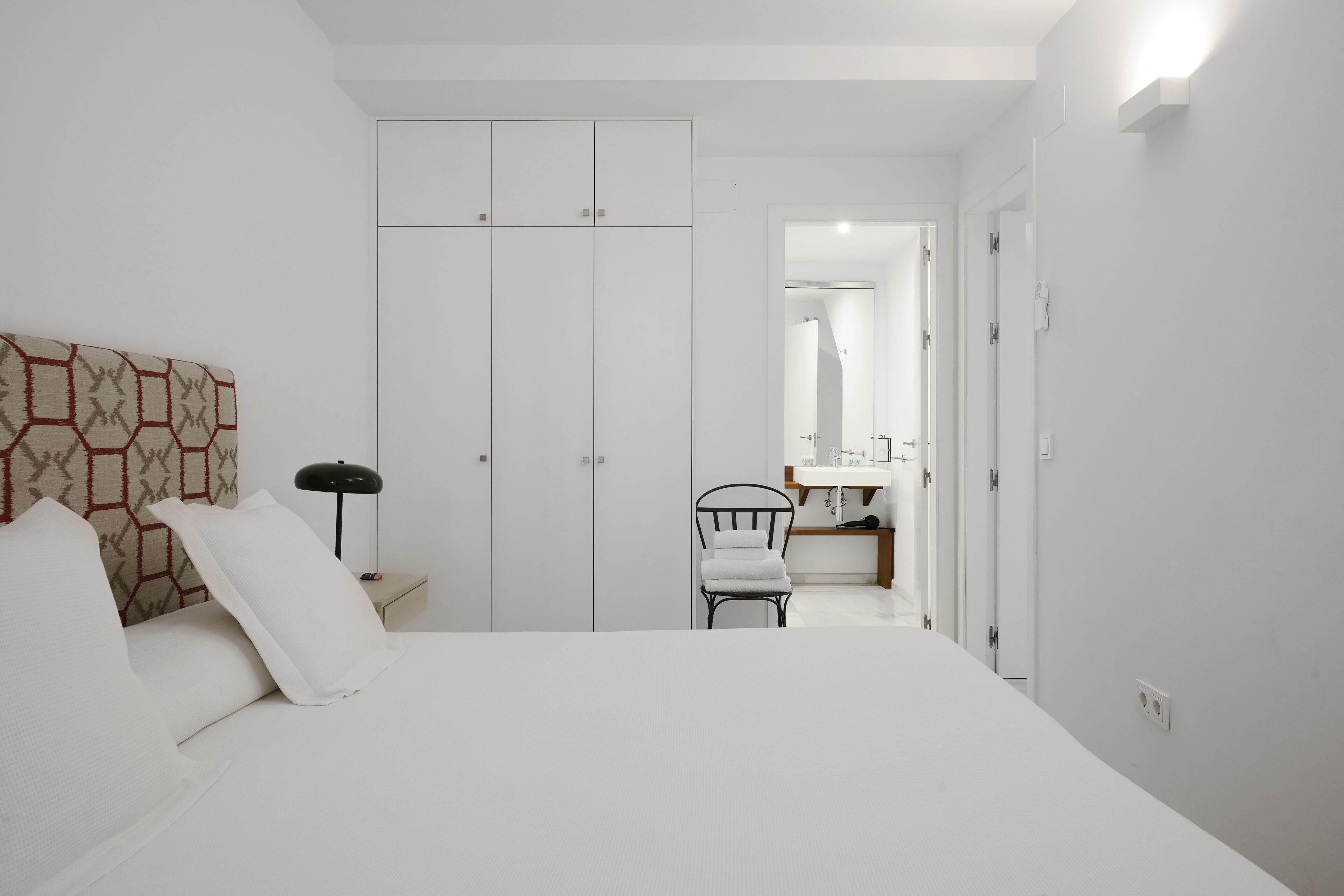 A bedroom with a bed, a nightstand, a chair, a wardrobe, and a bathroom visible through an open door.