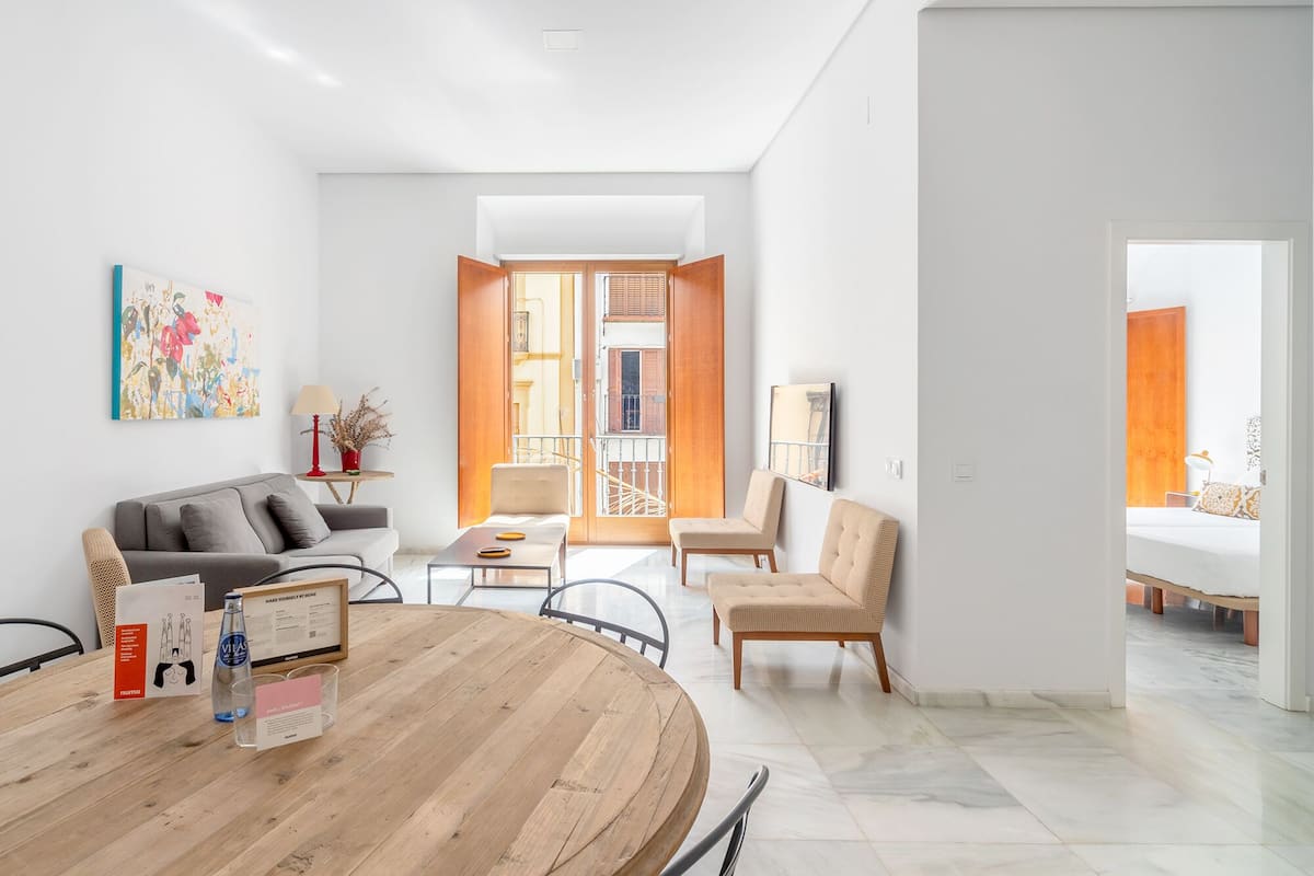 A modern living room with a round wooden table, a sofa, and two armchairs. There is a balcony with wooden shutters and a view of the outside.