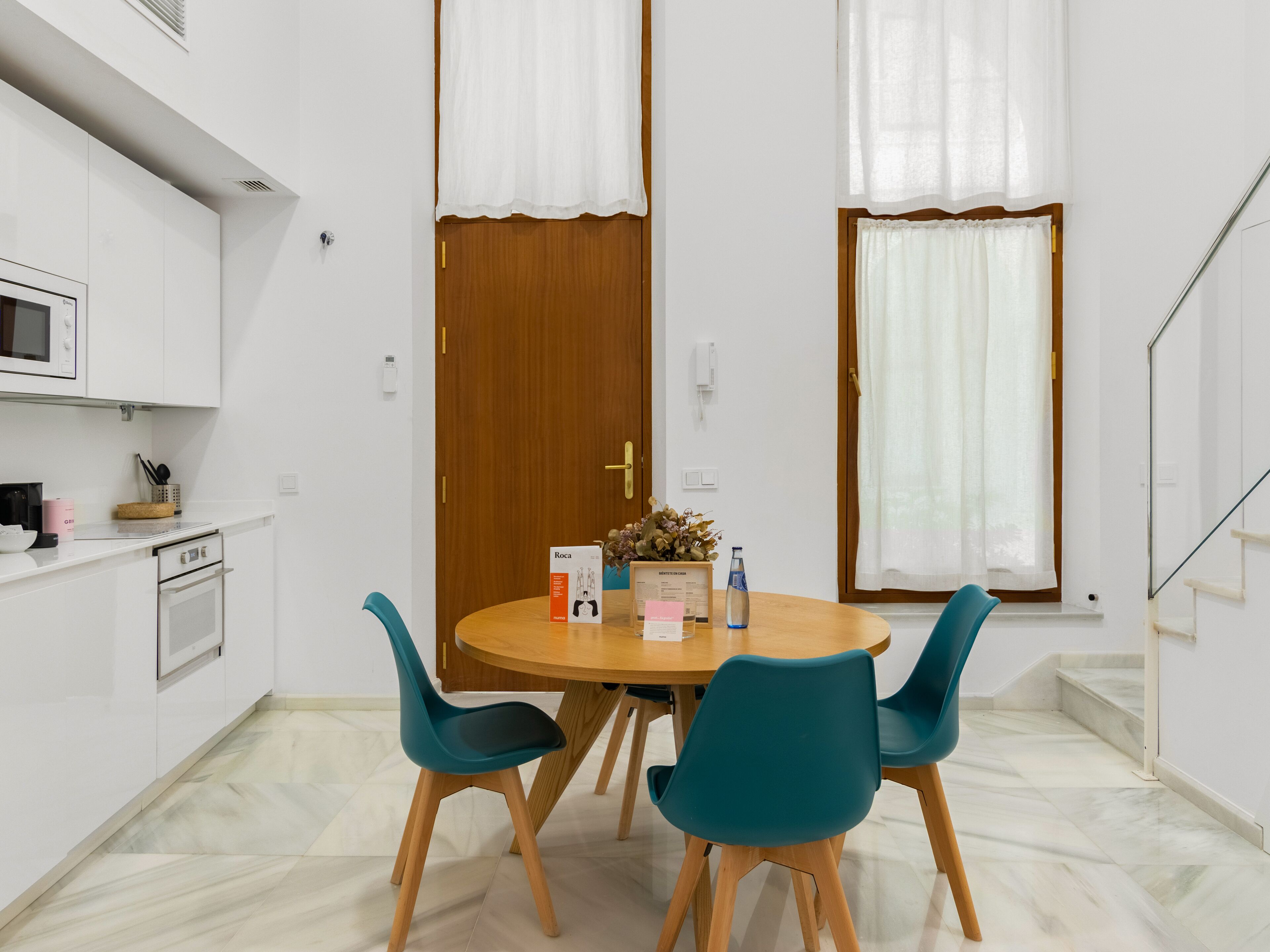 Una cocina moderna con armarios blancos, un microondas y un lavamanos. Hay una mesa de comedor de madera con dos sillas color turquesa y una pequeña planta en maceta.