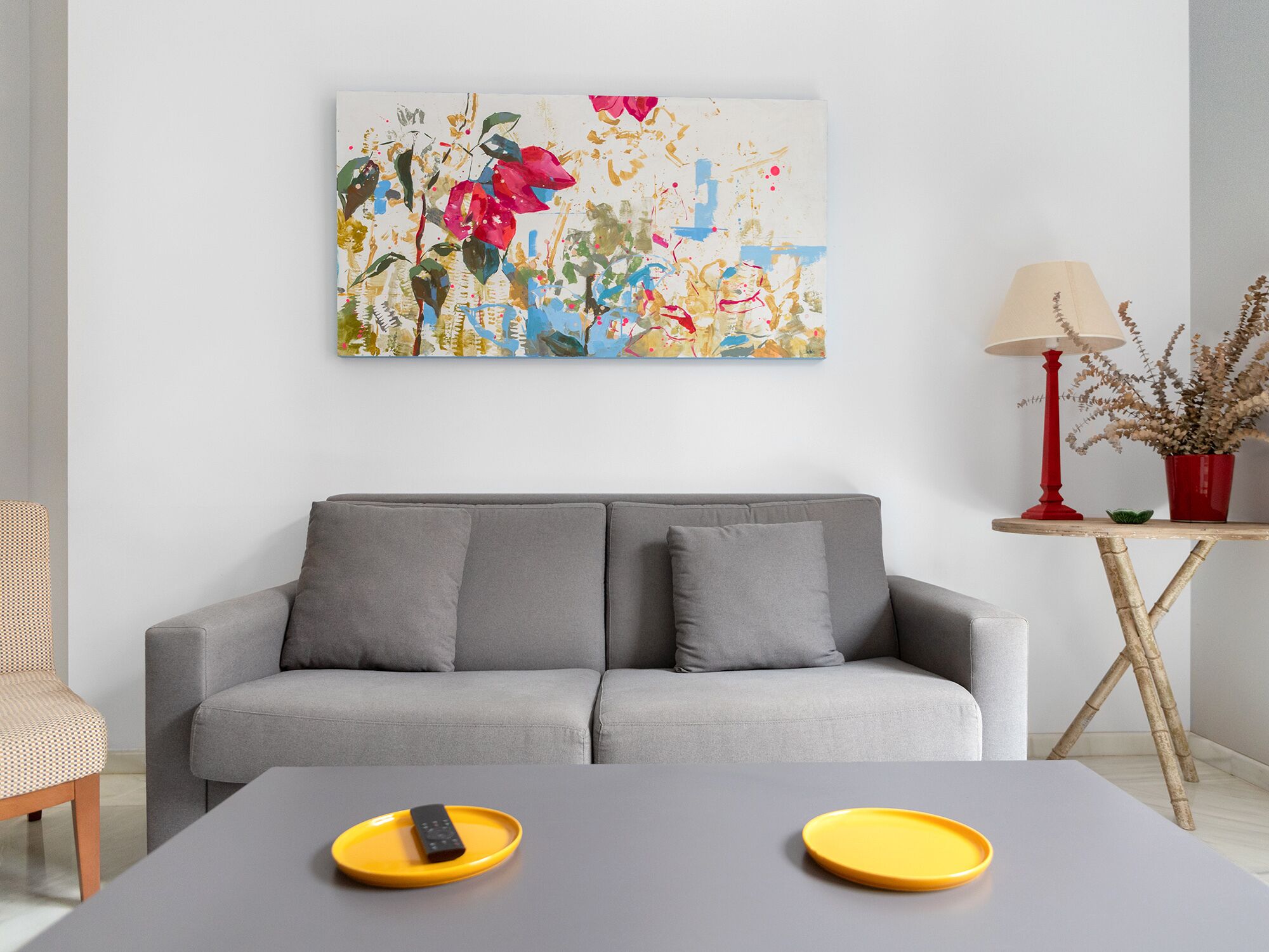 Ein modernes Wohnzimmer mit einem grauen Sofa, einem Couchtisch mit zwei gelben Tabletts, einer Fernbedienung, einer roten Lampe und einer Topfpflanze.