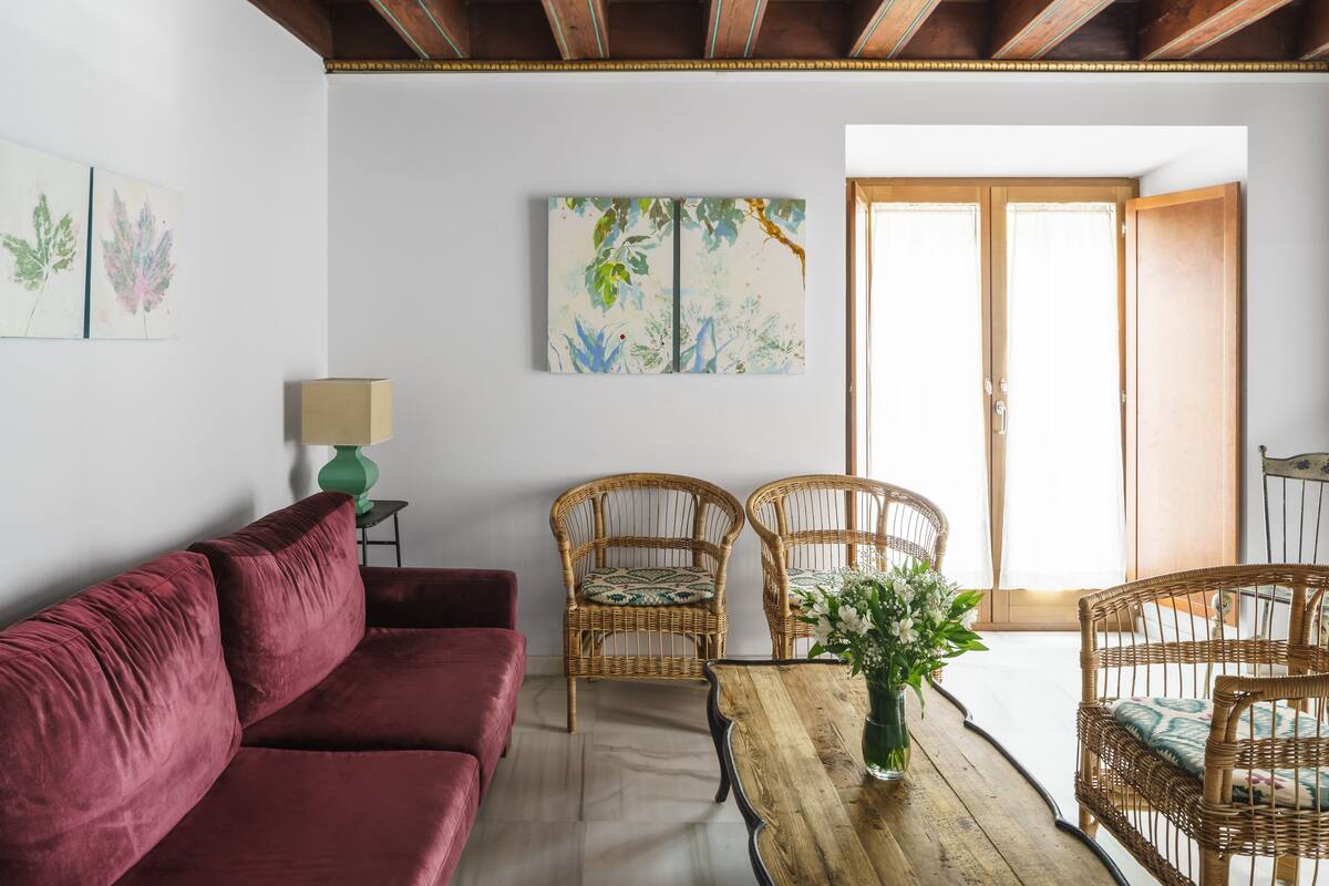 A living room with a red sofa, wicker chairs, a wooden table with a vase, and a painting on the wall.