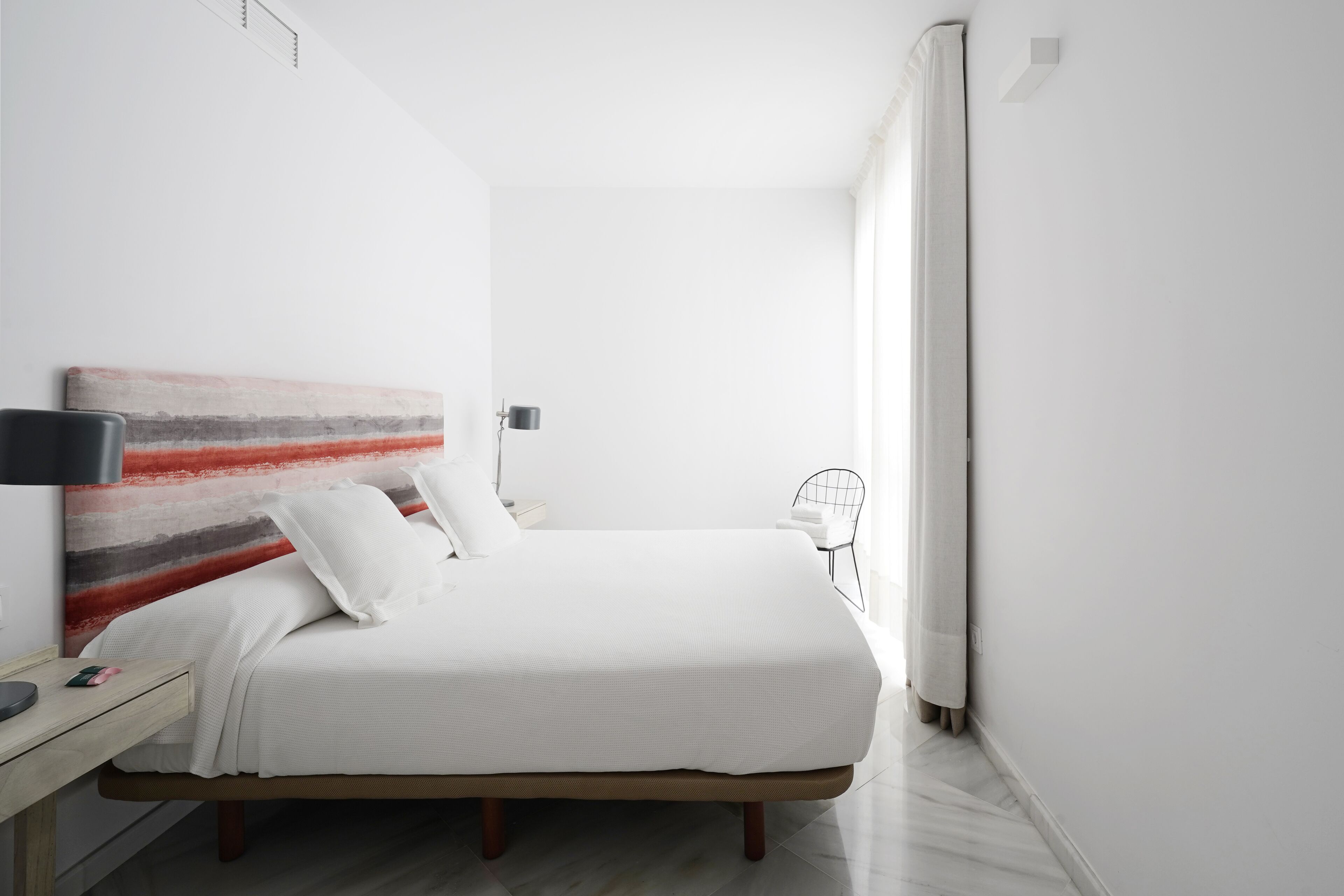 Un dormitori minimalista amb llit, taula de nit, làmpada i un quadre abstracte modern a la paret.