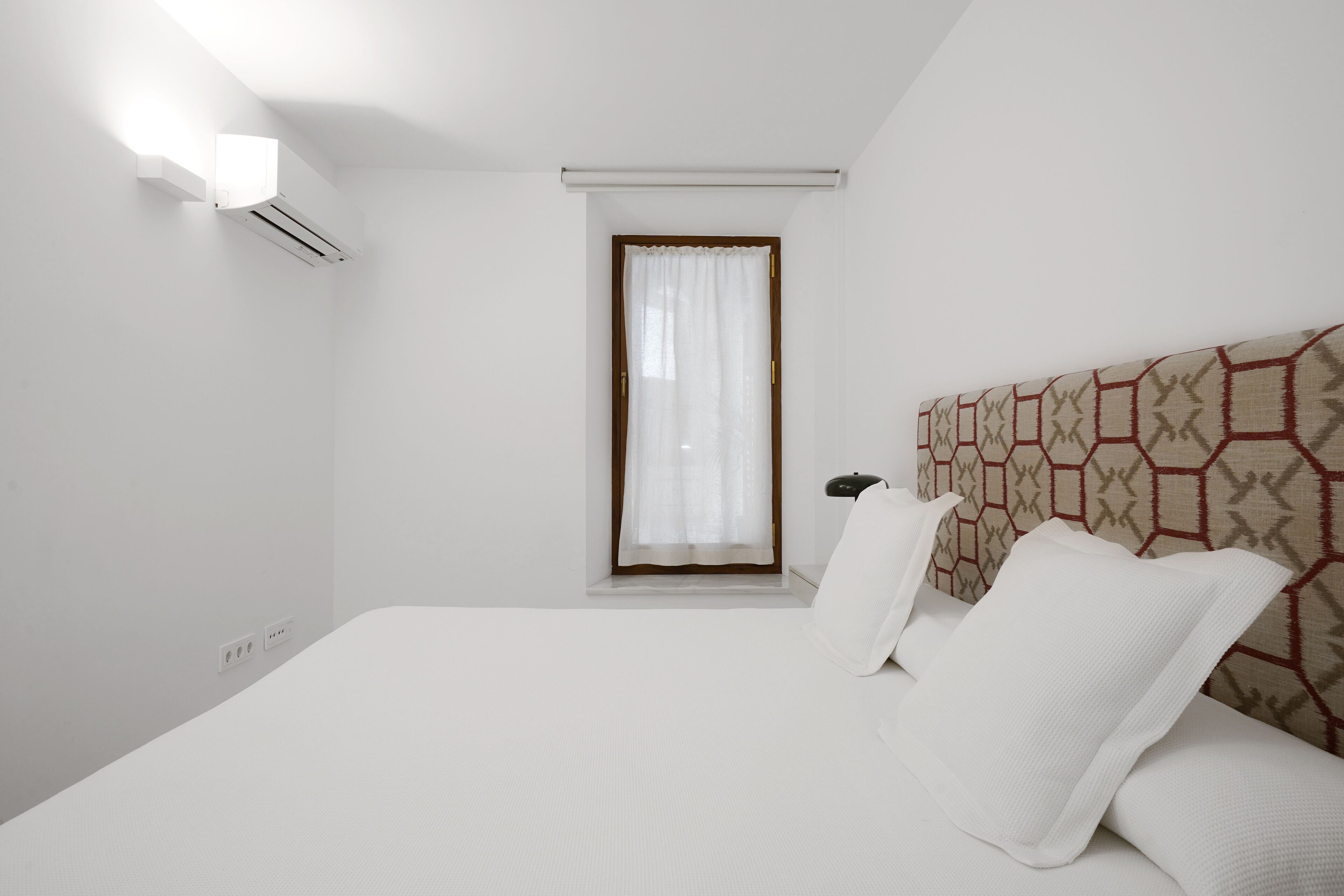 Una camera d'albergo con un letto grande, un comodino, una finestra con tende e un condizionatore d'aria a parete.