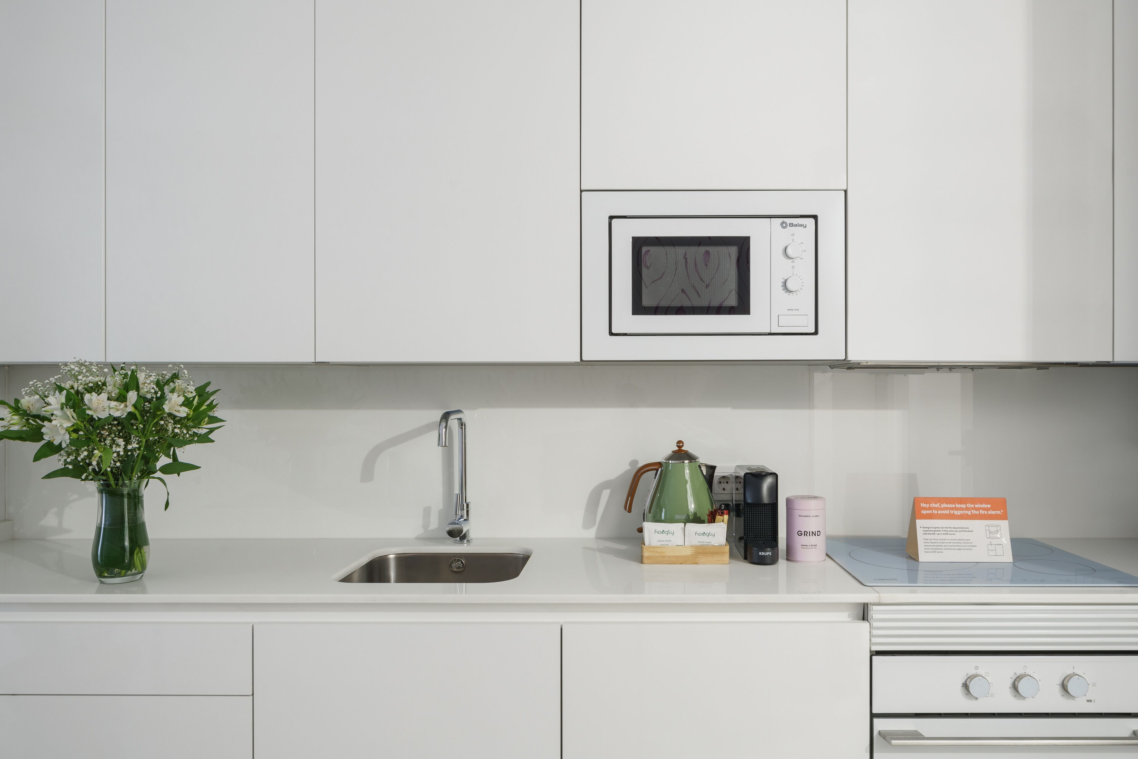 A modern kitchen with white cabinets, a stainless steel sink, a microwave, a kettle, a coffee maker, and a box of coffee pods.