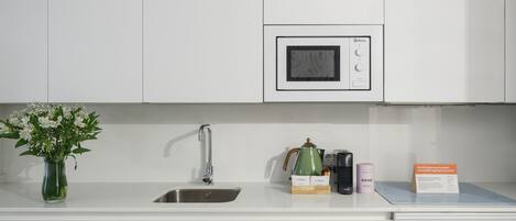 Eine moderne Küche mit weißen Schränken, einer Edelstahlspüle, einer Mikrowelle, einem Wasserkocher, einer Kaffeemaschine und einer Kapselbox.