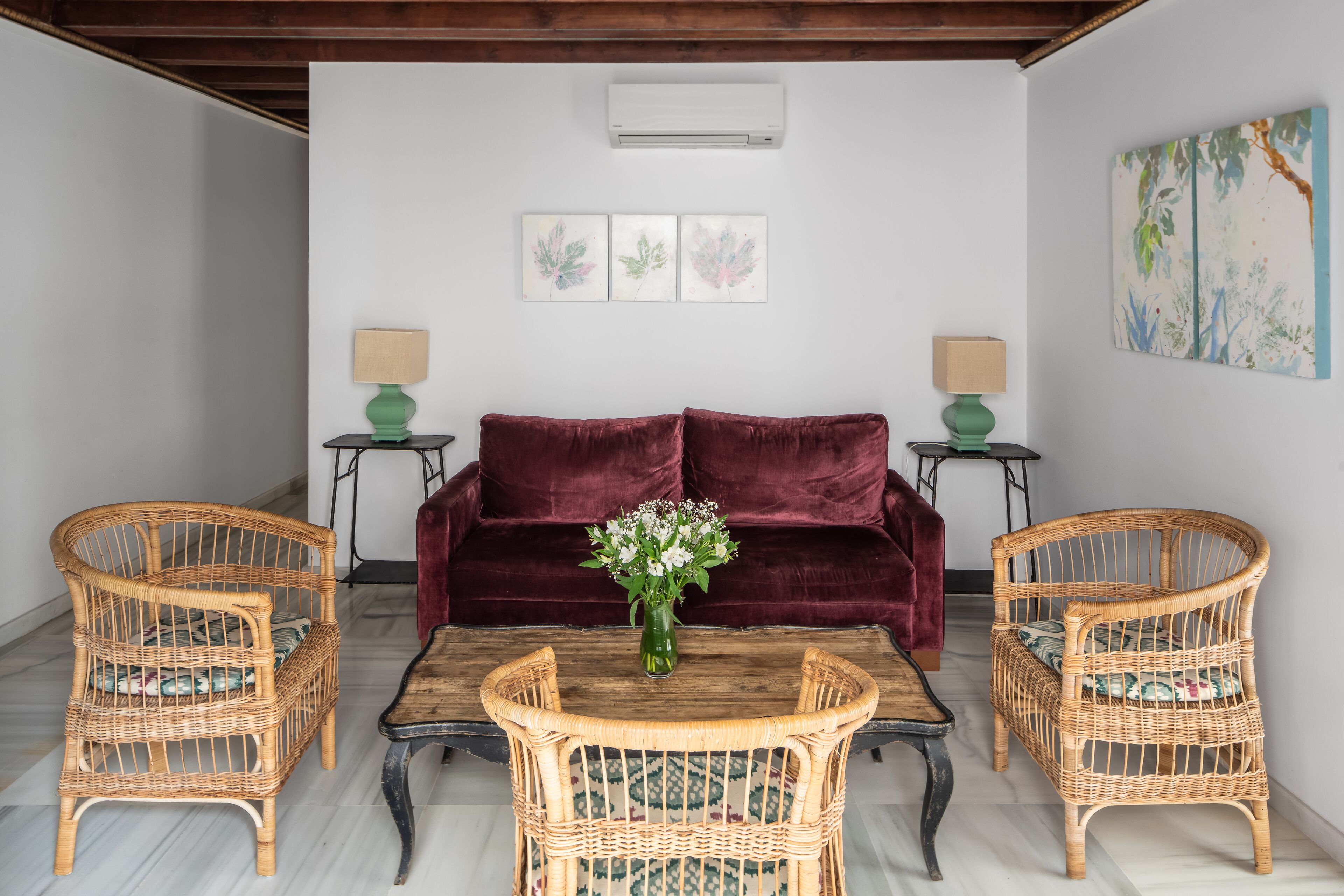 Un salon avec une table basse en bois, deux fauteuils en osier, un canapé en velours rouge et un climatiseur fixé au mur.