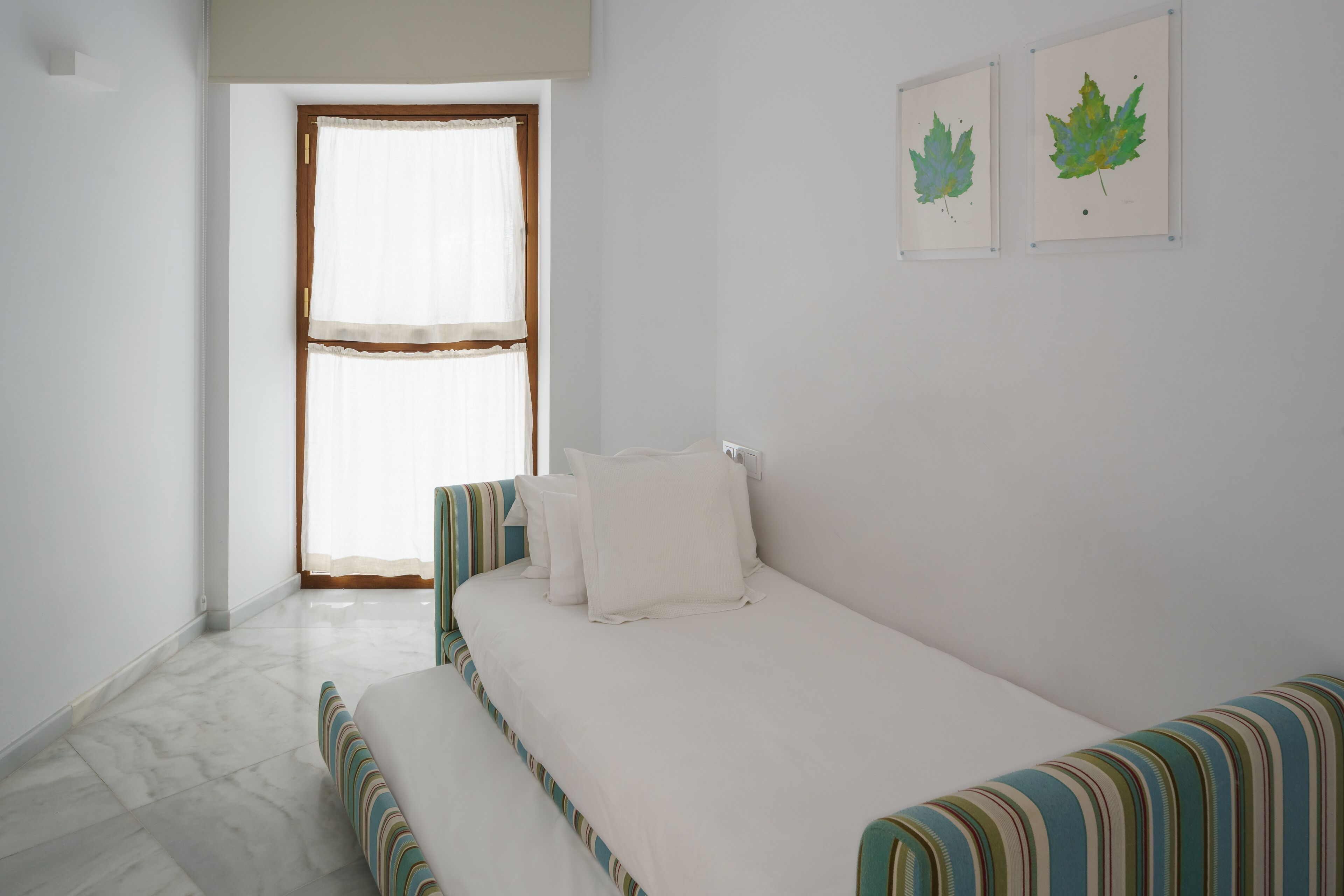 A bedroom with a bed, a bench, a window with curtains, and two framed leaves on the wall.