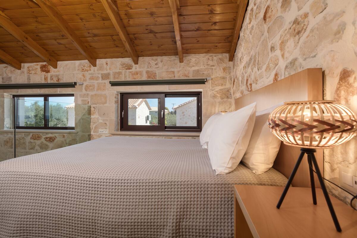 A bedroom with a stone wall, wooden ceiling beams, a bed with a white bedspread, a bedside lamp, and a window with a view of a house.