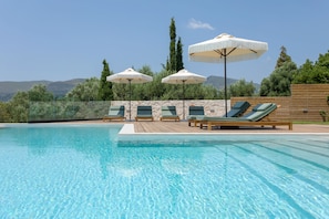 A swimming pool with lounge chairs and umbrellas, surrounded by a stone wall and greenery, with mountains in the background. - Evylio Stone Houses (Zakynthos)