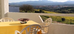 A balcony with a yellow table and white chairs, overlooking a vineyard and distant mountains. - B&B Anacleto (Luogosanto)