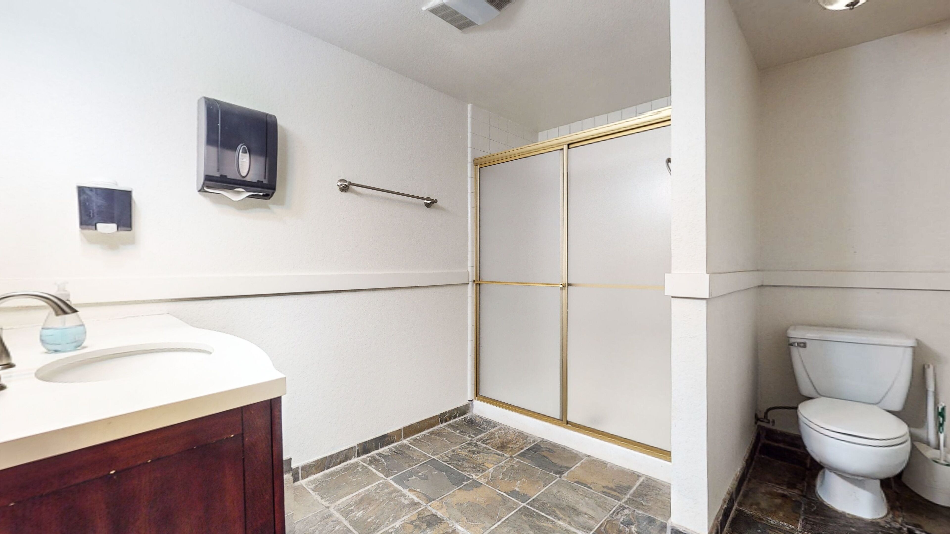 A bathroom with a shower, toilet, sink, and a soap dispenser on the wall.