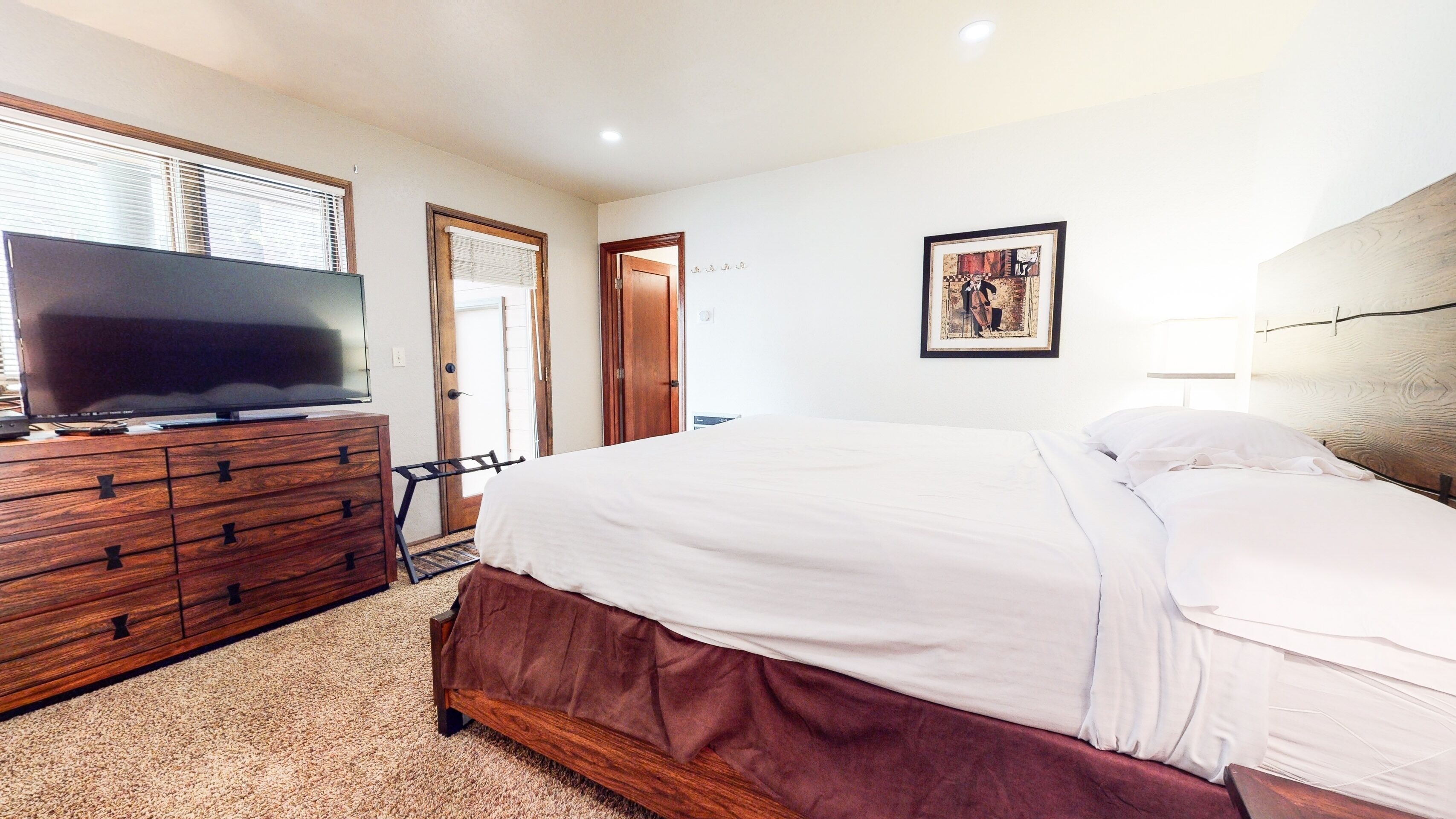 Un dormitorio con cómoda de madera, televisor de pantalla plana, cama con ropa de cama blanca, una puerta y un cuadro en la pared.