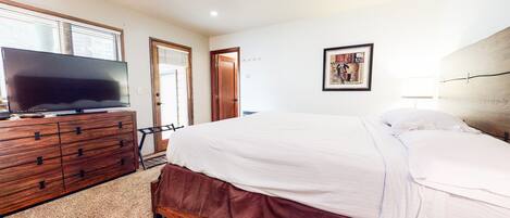 Un dormitorio con cómoda de madera, televisor de pantalla plana, cama con ropa de cama blanca, una puerta y un cuadro en la pared.