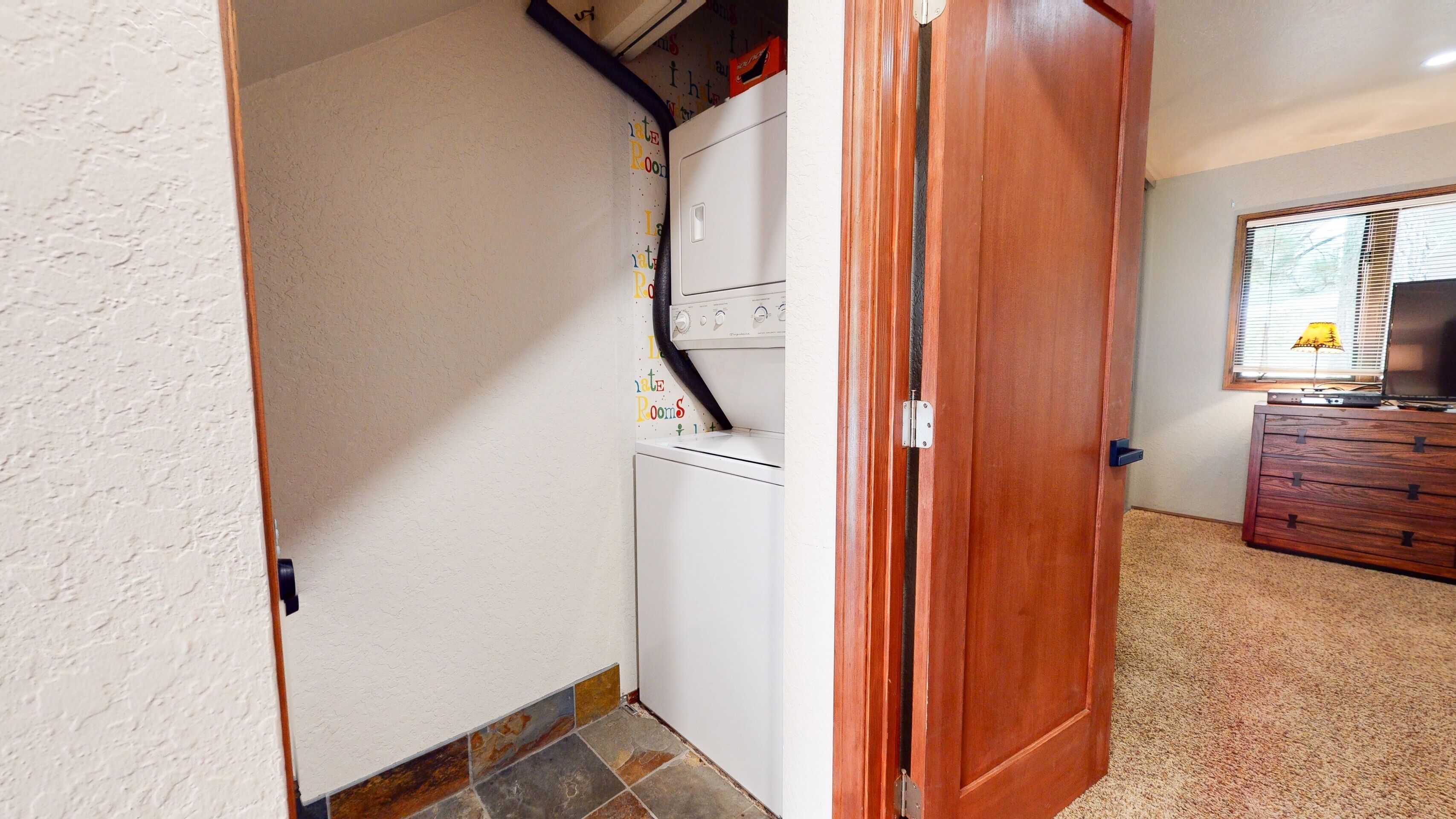 A laundry room with a washing machine and dryer, a wooden door, a dresser, and a window with blinds.
