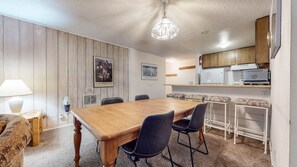 A dining area with a wooden table and chairs, a kitchen with appliances, and a ceiling light fixture. - Summit #273 2 Bedroom Condo by RedAwning (Mammoth Lakes)