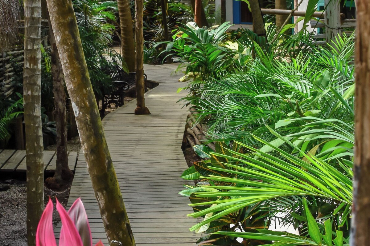 A wooden pathway surrounded by lush tropical plants and palm trees, leading to a thatched-roof structure.