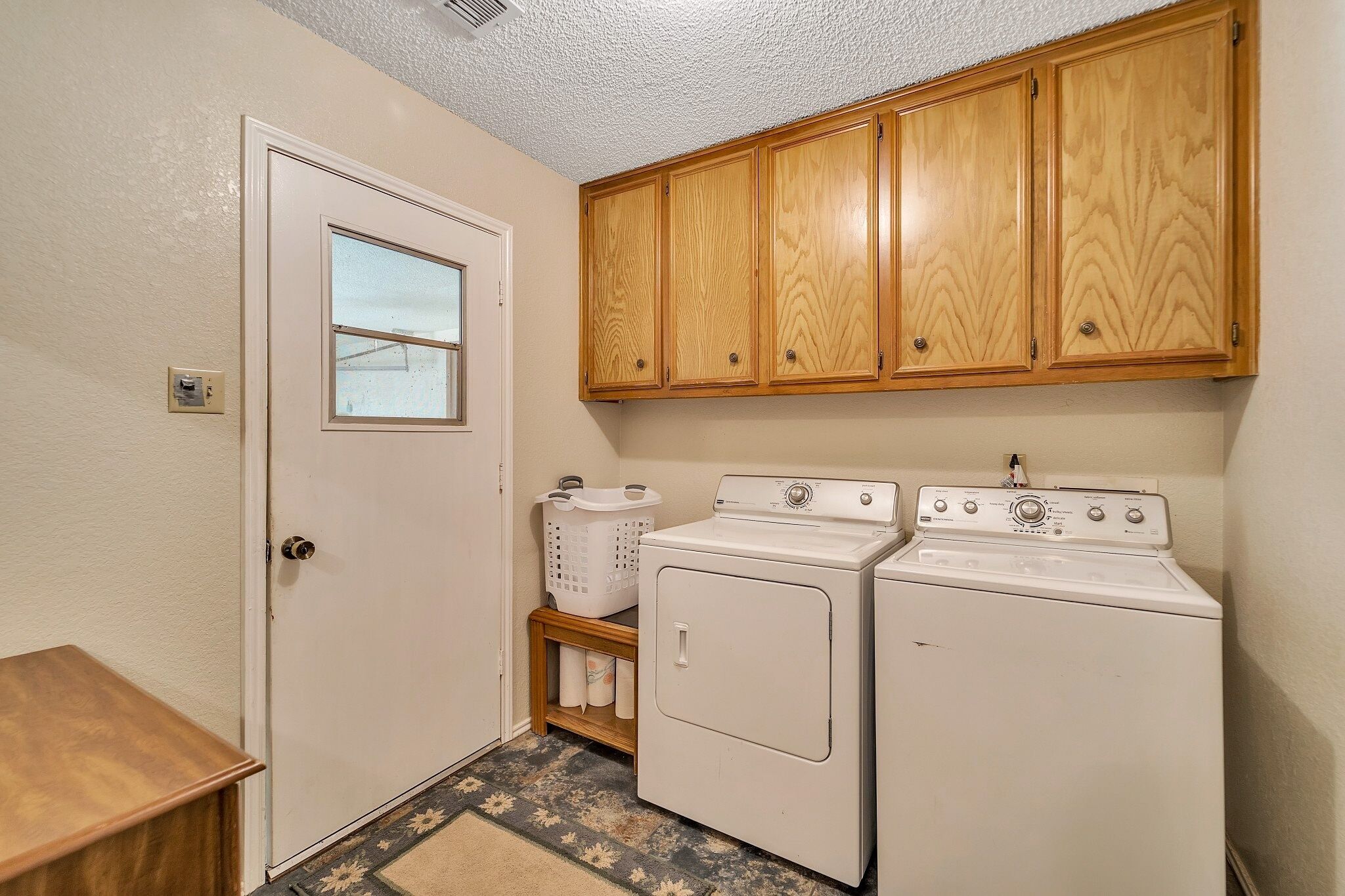 A laundry room with wooden cabinets, a washing machine, a dryer, a door with a window, and a small shelf with laundry supplies.