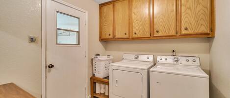 A laundry room with wooden cabinets, a washing machine, a dryer, a door with a window, and a small shelf with laundry supplies.