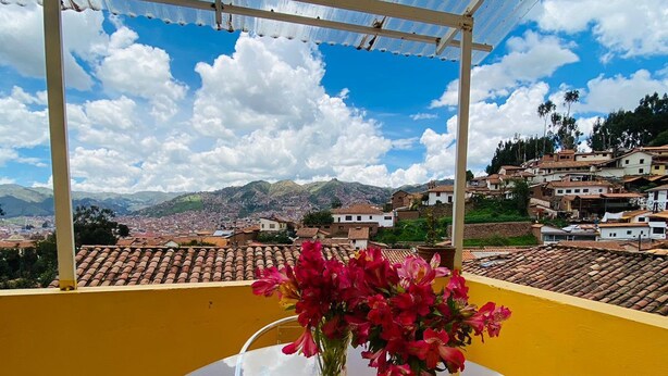 A glass table with chairs and a vase of flowers on a balcony with a yellow wall and a view of a town with terracotta roofs and mountains in the background. - Apartments Stunning View San Blas (Cusco)
