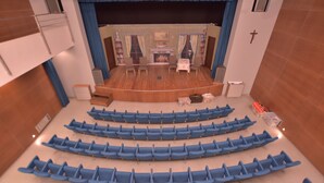 Un teatro con hileras de asientos azules, un escenario con chimenea y una cruz en la pared.