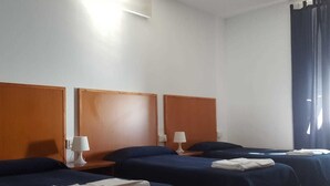Habitación de hotel con tres camas individuales, cabeceros de madera, mesitas de noche con lámparas y una ventana con cortinas oscuras.