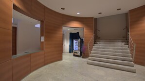 Un vestíbulo de hotel moderno con una pared curva de madera, piso de mármol, una escalera con barandales de metal y una máquina expendedora.