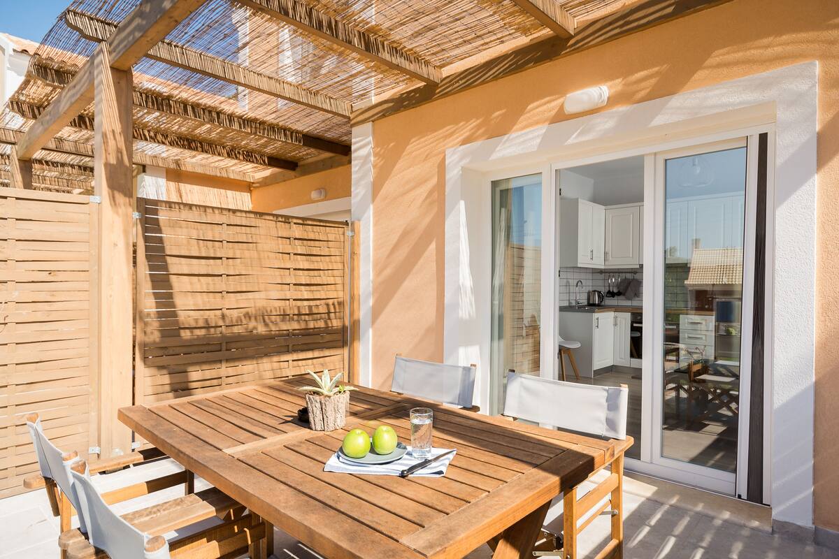 Een houten tafel met stoelen, een glas water en twee appels op een terras met een houten hek en een schuifdeur naar een keuken.